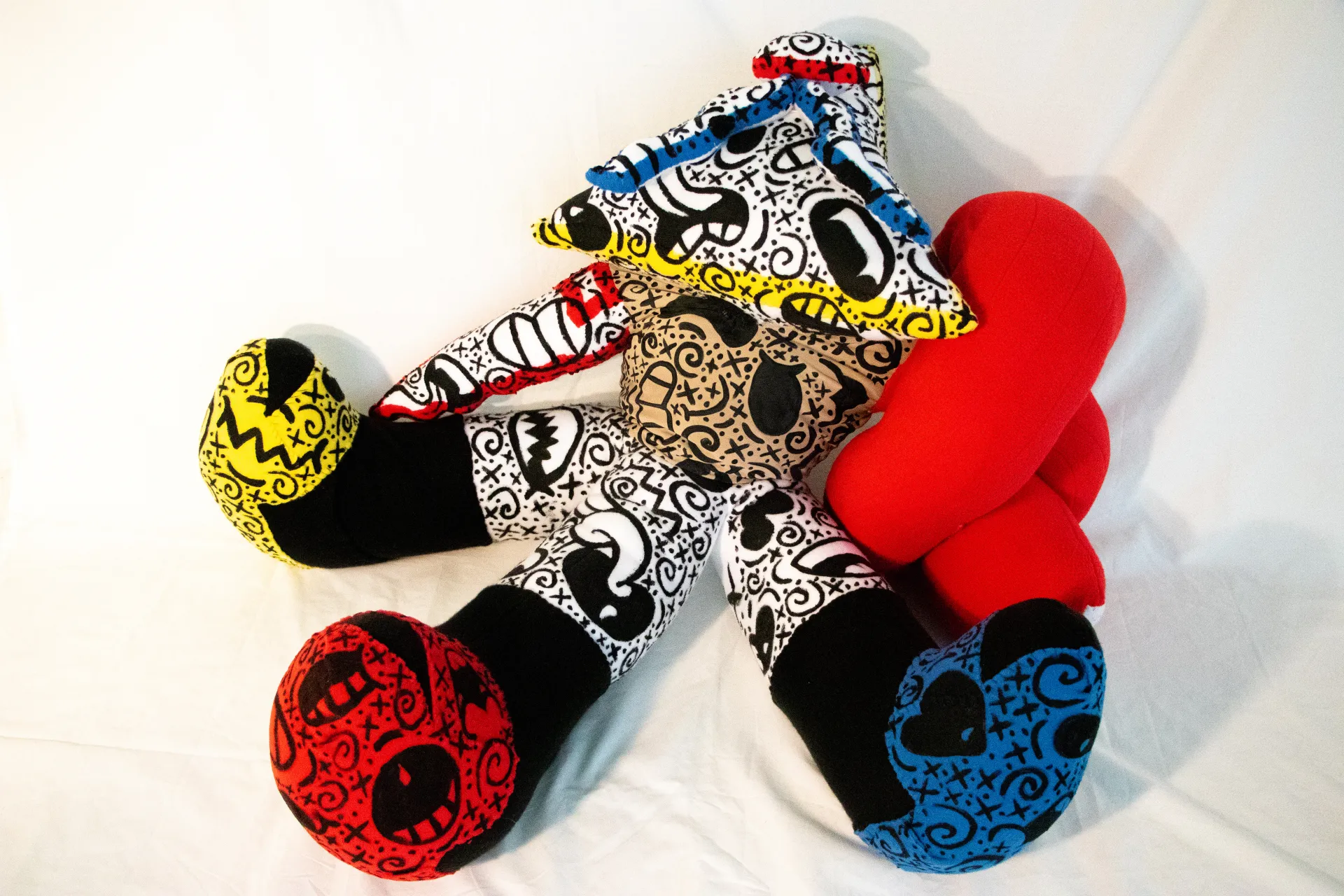 A photograph of a fabric sculpture in bright red, yellow and blue with multiple(3) appendages and covered with swirling black markings, save for one purely red portion and three entirely black pieces near the end of the appendages.. 