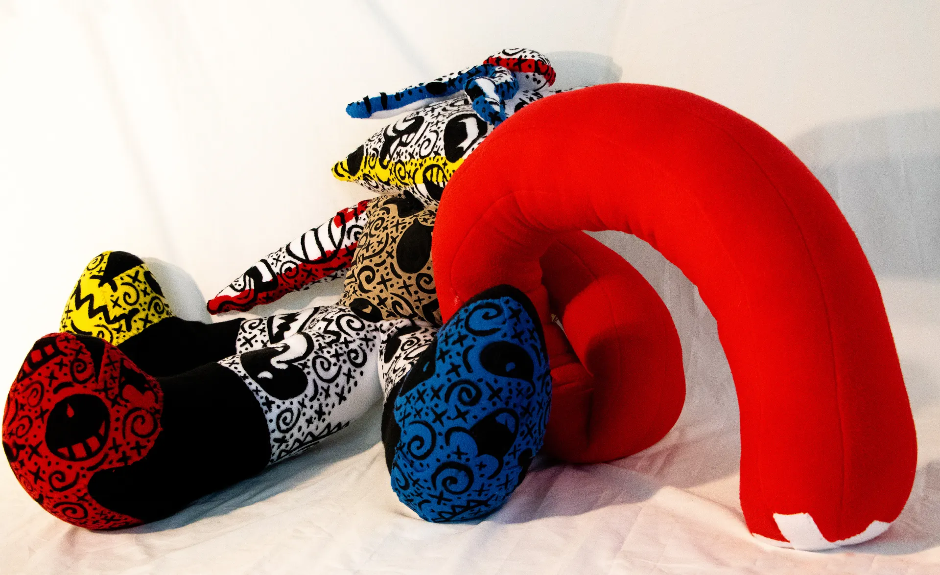A photograph of a fabric sculpture in bright red, yellow and blue with multiple appendages and covered with black markings. Here the solely red piece is arched from the central form.