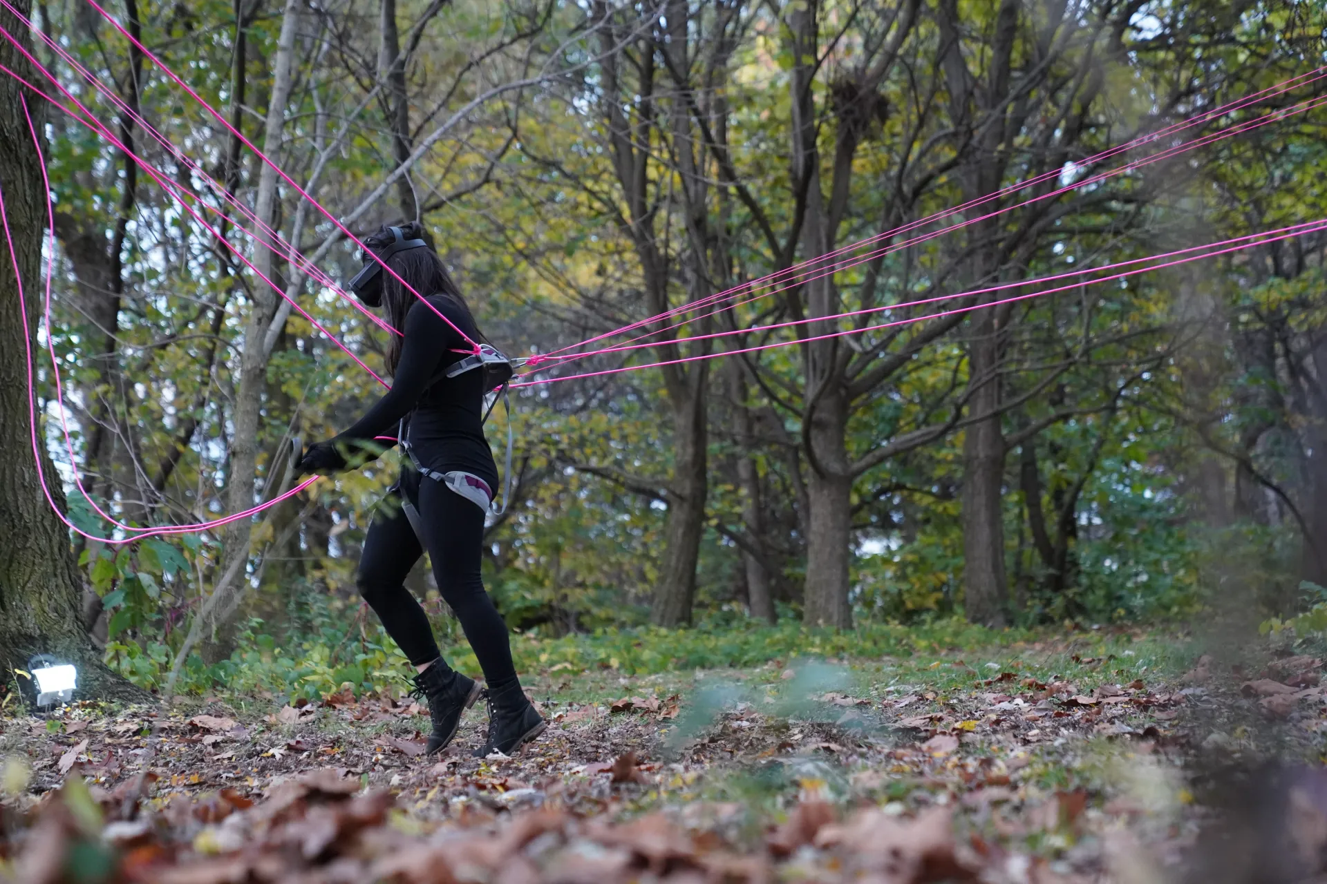 A close up of the figure, wearing all black and a VR headset, in action, attached to the trees by neon pink string.