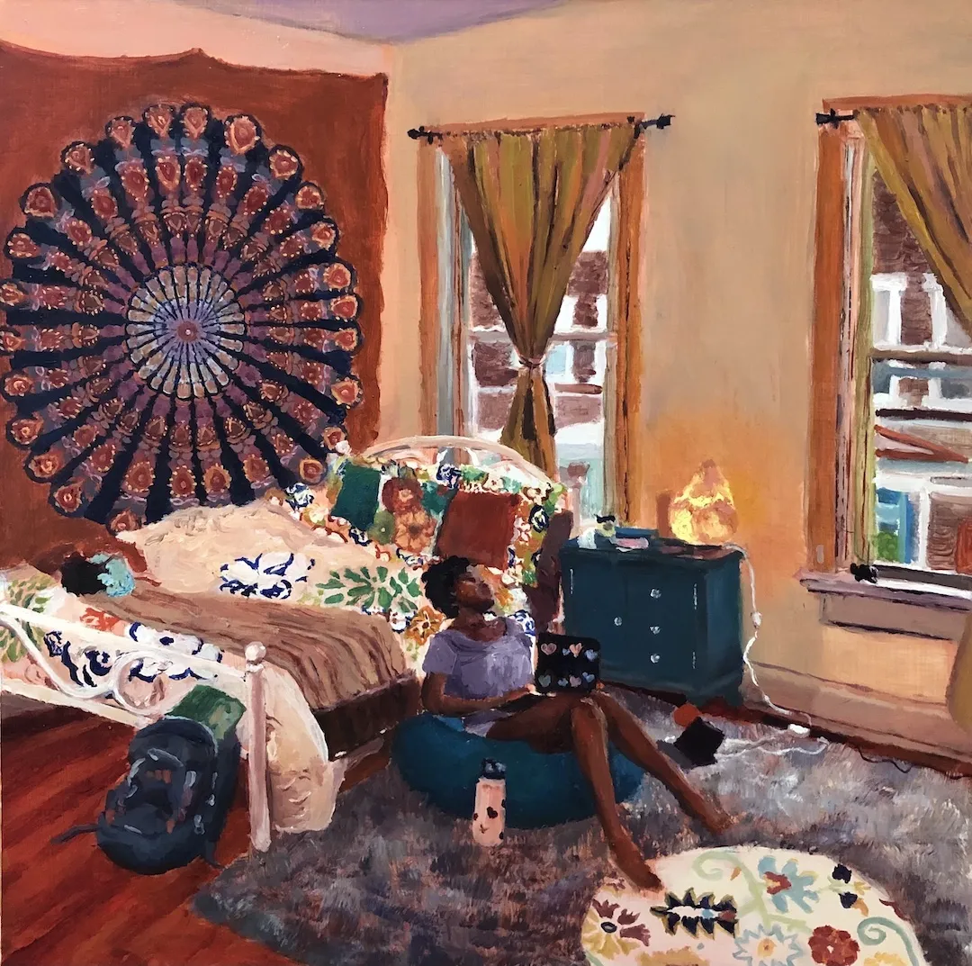 A person sits on a cushion, holding a laptop, on the floor of a warmly lit bedroom