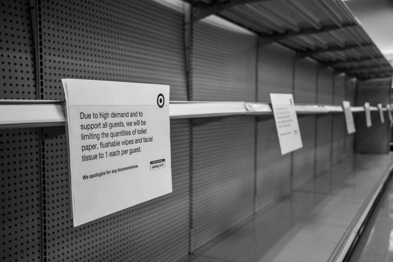 A black-and-white photo of empty store shelves with signs informing guests the amount of paper products they can buy are limited