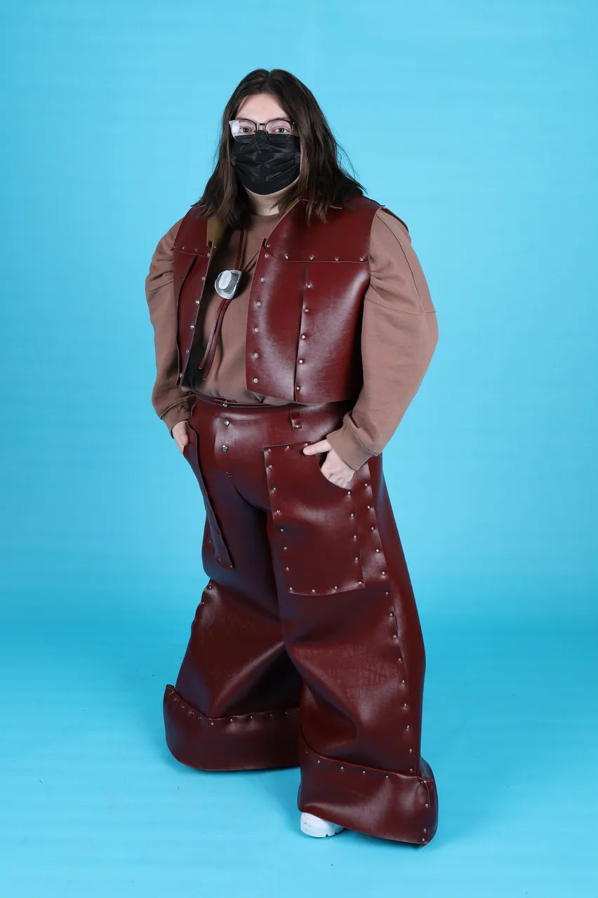 A model wears reddish-brown vinyl pants and vest over a light brown long-sleeve shirt; they also wear a bolo tie with a cowboy hat-shaped clasp.