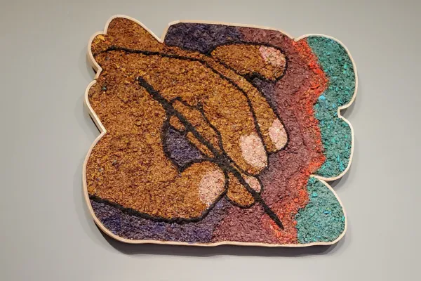 A painting, made of arranged and colored wood chips, depicts a hand holding a writing utensil in front of a red, green, and purple background.