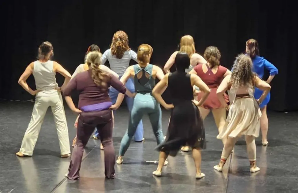 ten dancers stand with wide legs, hands on hips, and backs facing camera