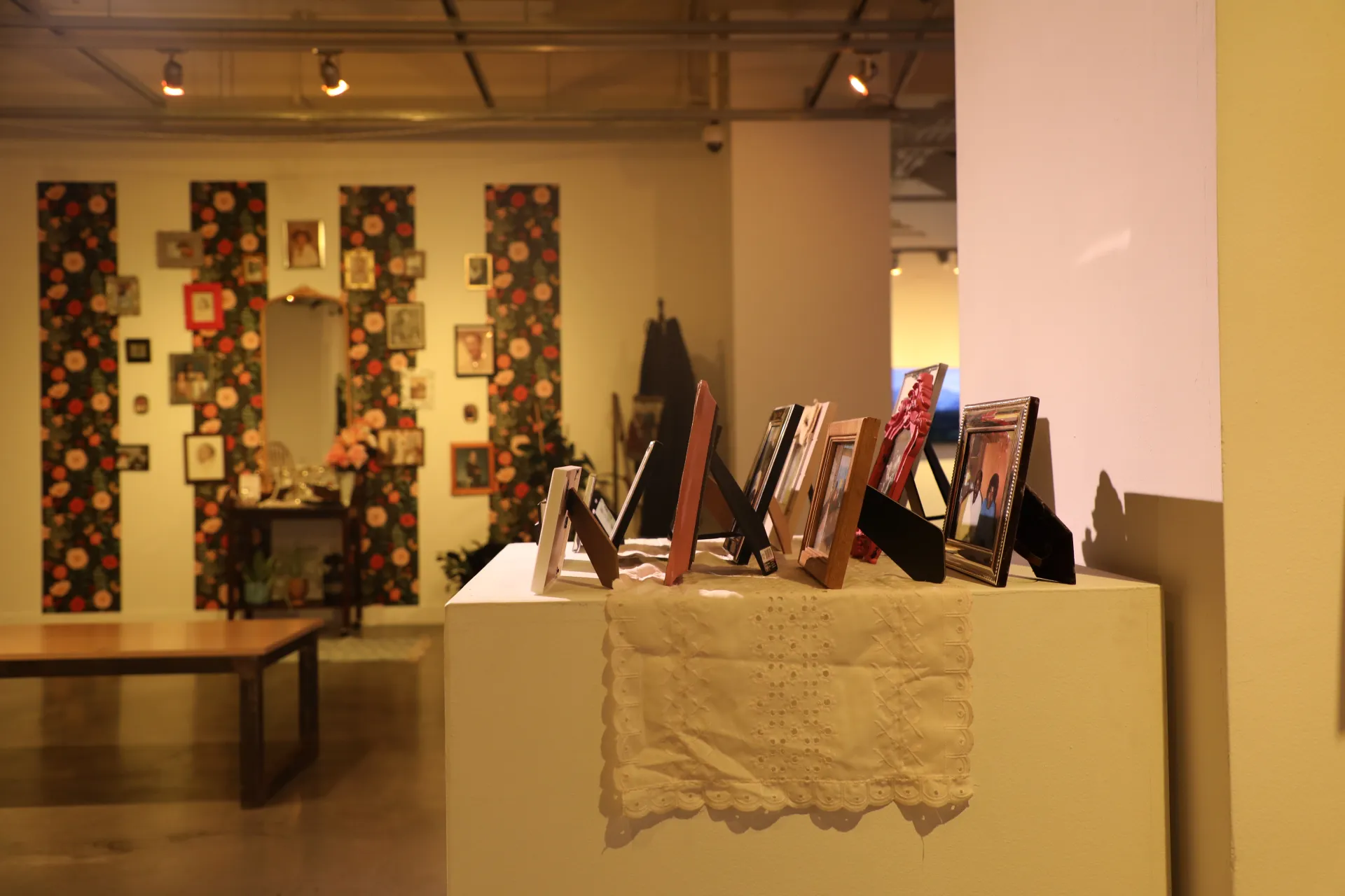 Around 10 framed photographs sit on a lace doily. In the background, a mirror hangs above a desk with framed photos on the wall and flowery wallpaper