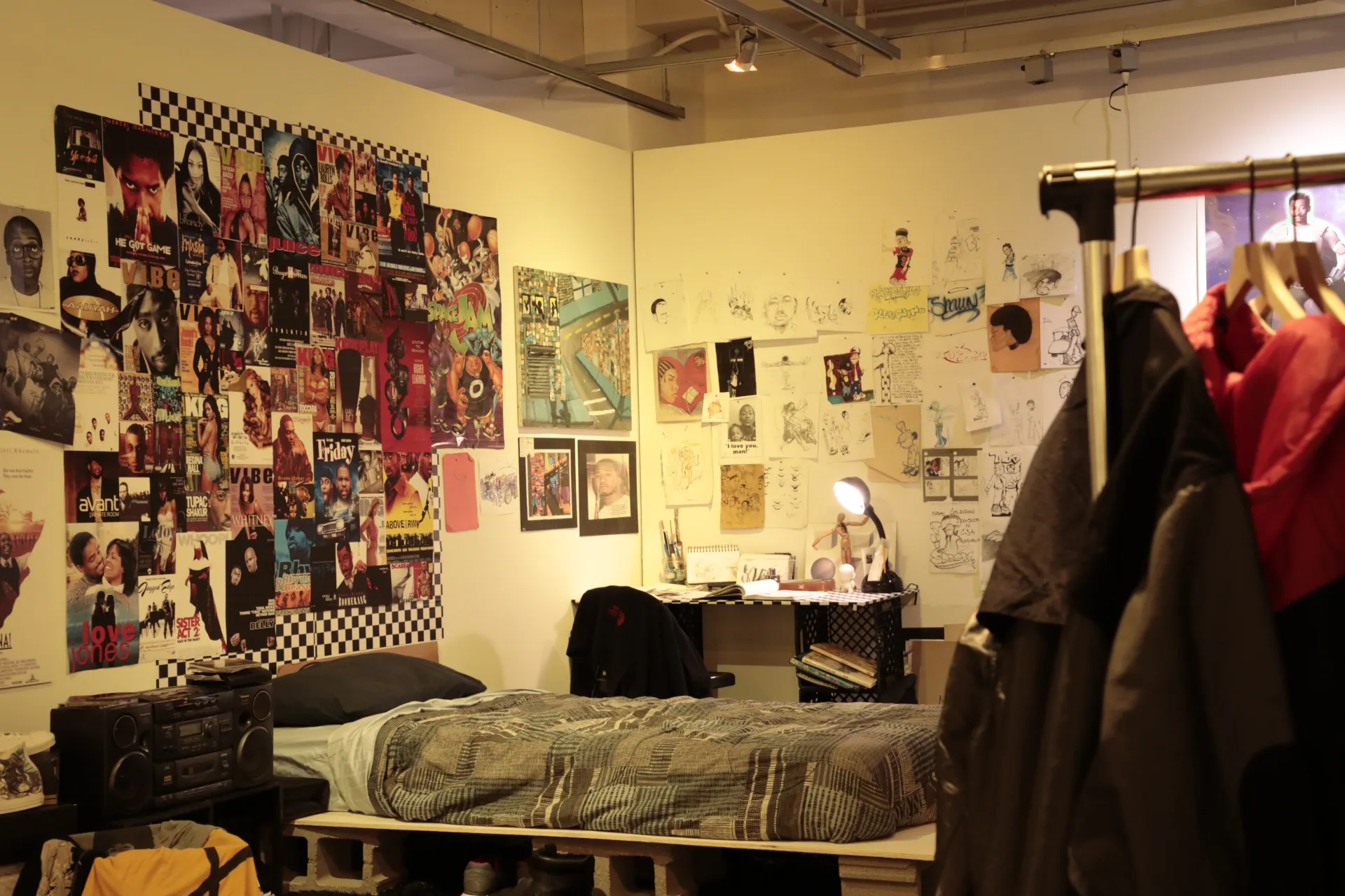 Bedroom with a collage of posters and magazine covers above the bed. Behind the bed is a desk with drawings on the wall. A coat rack with jackets is to the right. 