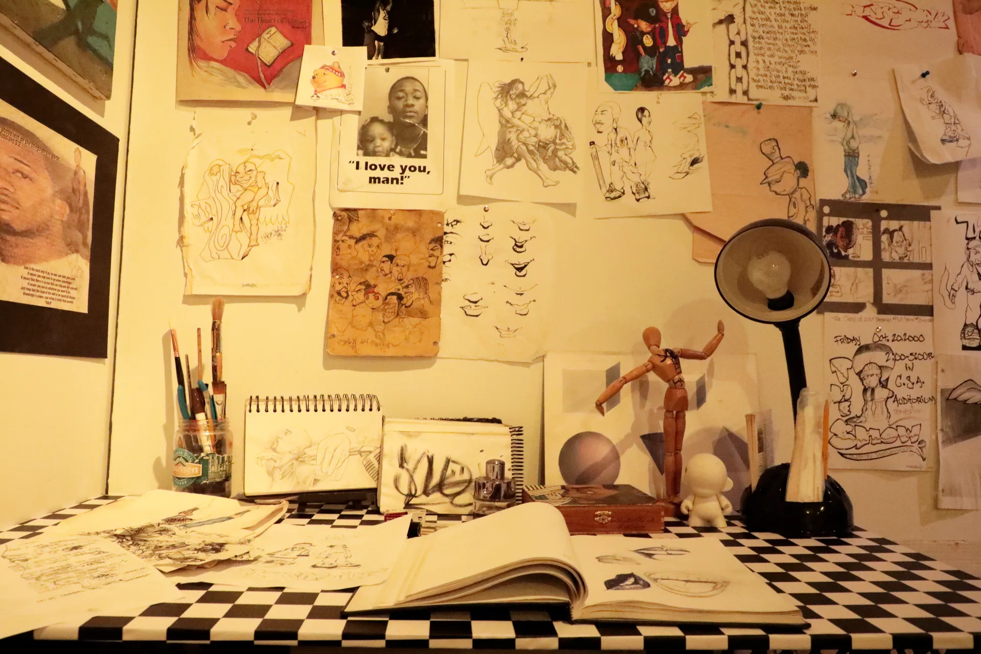 A checkerboard desk with an open sketchbook and various other pages with illustrations. A lamp and wooden drawing mannequin sit to the right, with pencils and pens to the left. The wall is covered in drawings. 
