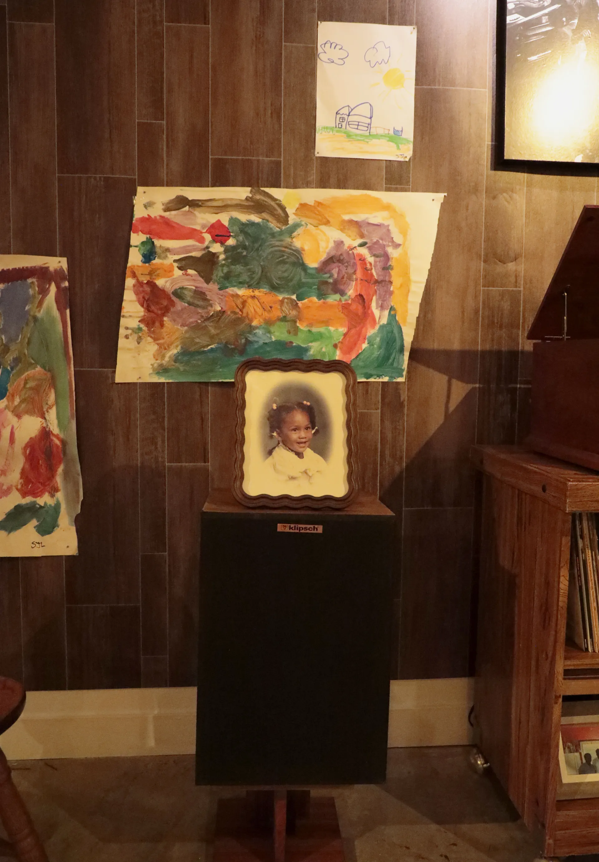 A framed photograph of a girl sits on top of a speaker. On the wall behind the speaker is children's art work and paintings. 
