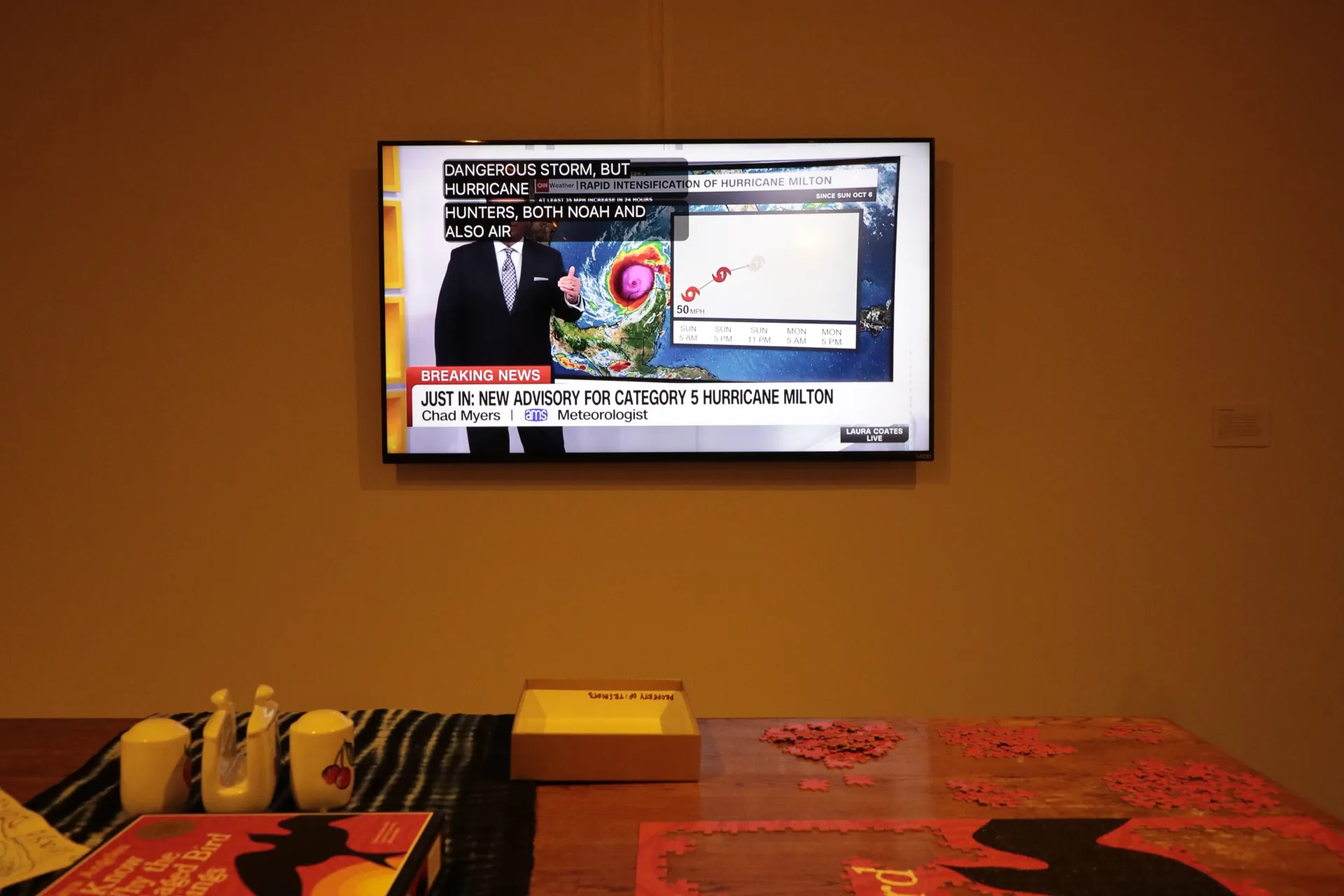 A table with a puzzle in progress. Above the table is a TV playing the weather channel, reporting on a hurricane advisory 