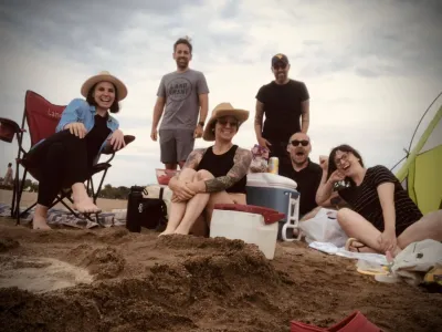 A group of six people sitting on the beach and smiling at the camera surrounded by coolers and blankets