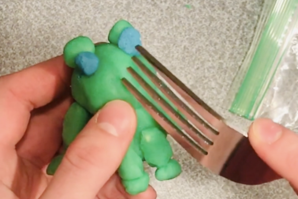 still from video, a clay frog is being held as a fork is used to create the imprint of a nose