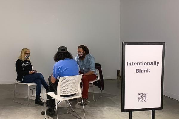 Three people sitting at a table. A sign in front of them says "Intentionally Blank" 