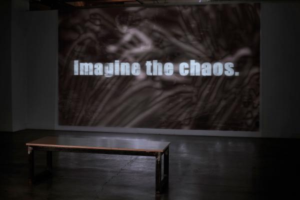 a wall with a video projected onto it with a bench nearby, projected video still says "imagine the chaos"