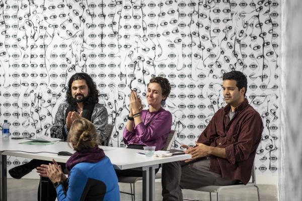 Front view of three artist panelists, Illya Mousavijad, Fatemeh Shams, and Omid Shekari sitting at a table While Dr. Merijn crouches in front of the microphone on the table to ask a question. On the wall behind them is Parastou Forouhar’s piece The Eyes, a large digital print of a mass of eyes with figures emerging between them. 