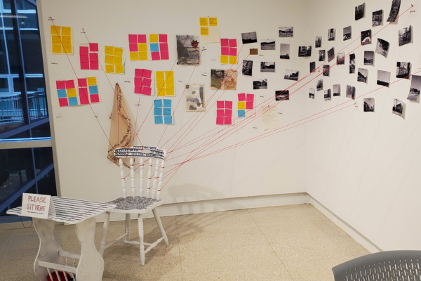 A white chair sits facing away from the corner of the gallery. Red strings attached to the back of the chair are connected to the wall. On the wall are multicolored sticky notes and black-and-white photos.