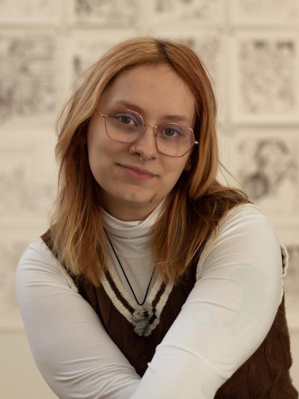 A person with light-brown hair, glasses, and a septum piercing smiles softly at the camera.