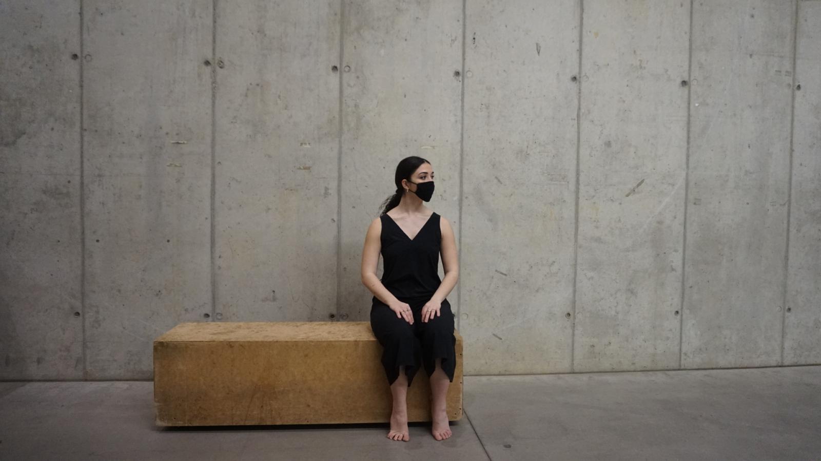 girl wearing mask, sitting on a bench in a room made out of concrete