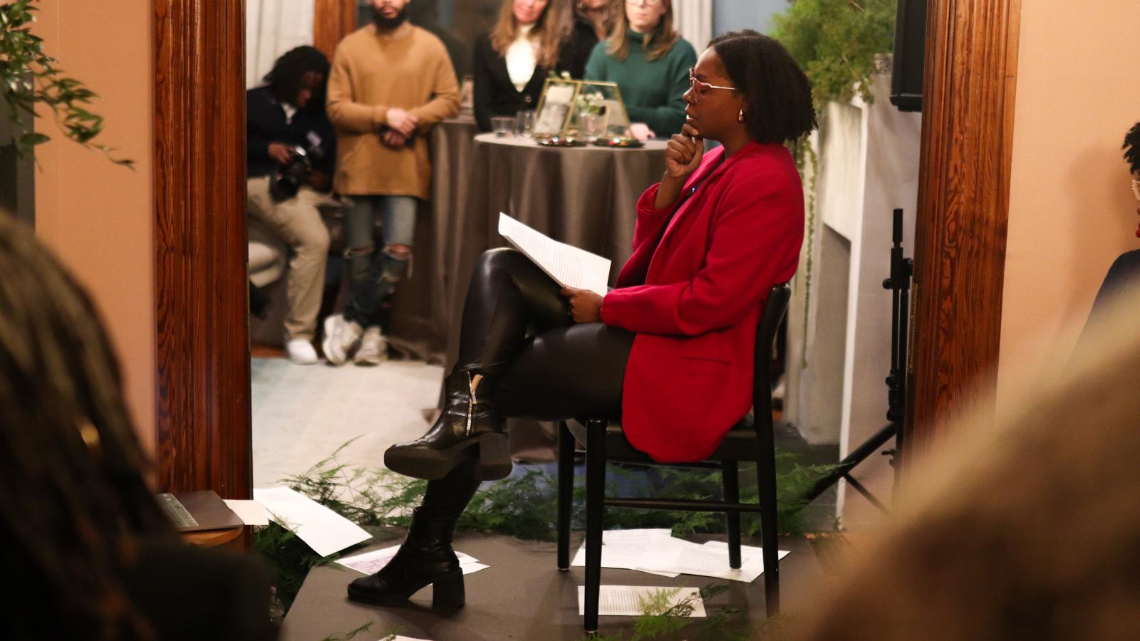 Ajanae in a red suit jacket sitting in a chair and performing poetry