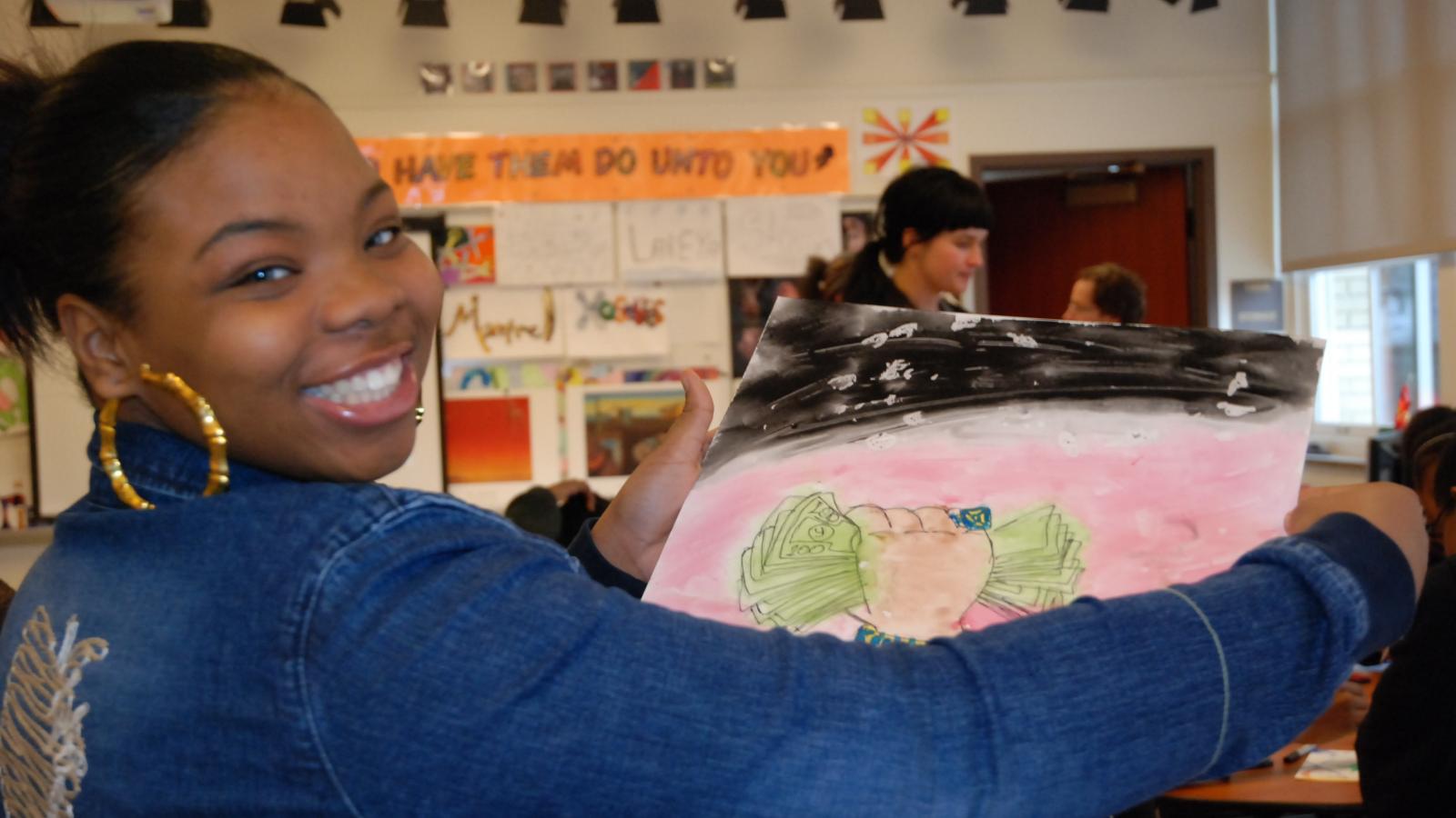 A young person smiles and holds up a piece of artwork