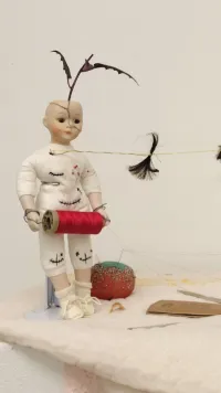 A close-up view of the assemblage. The baby doll covered in black stitches holds a spool of red thread. Its face is cracked and pins are stuck in its body.